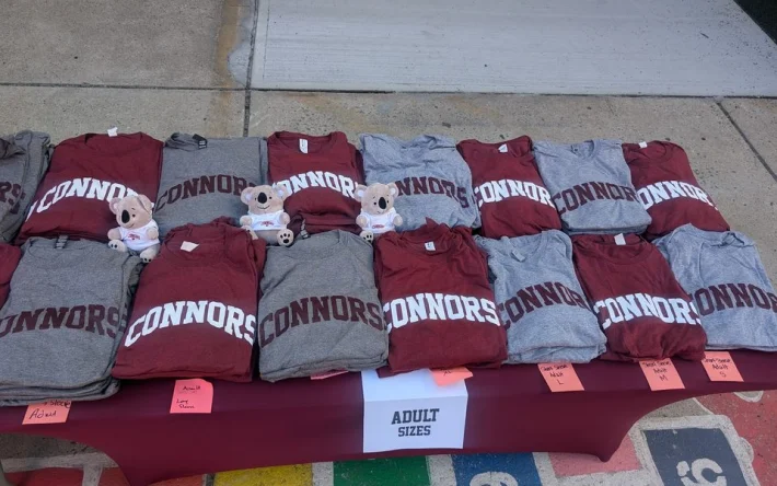 Folded maroon and grey Connors tees laid out with plush Koalas beside an Adult Sizes sign