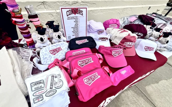 A wide spread of Connors gear: pink belt bags, visors, ball caps, bucket hats, sticker sheets, and plush Koalas