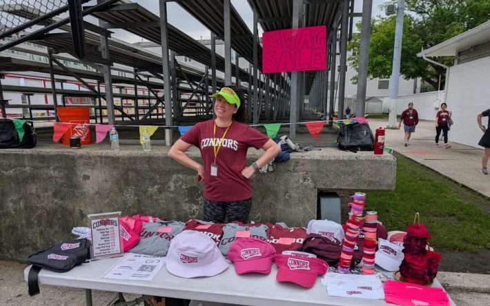 A Connors parent standing behind a swag sale table of tees, caps, and visors under a Swag Sale sign