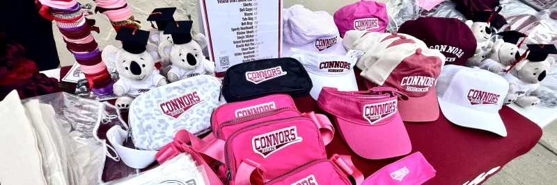 A Connors swag table spread with pink belt bags, visors, ball caps, and plush graduate Koalas