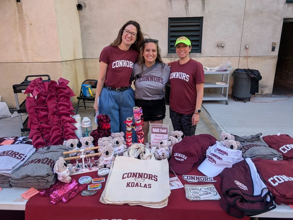 Three Swag Team members behind a full swag table of tees, tote bags, plush Koalas, and hair bows