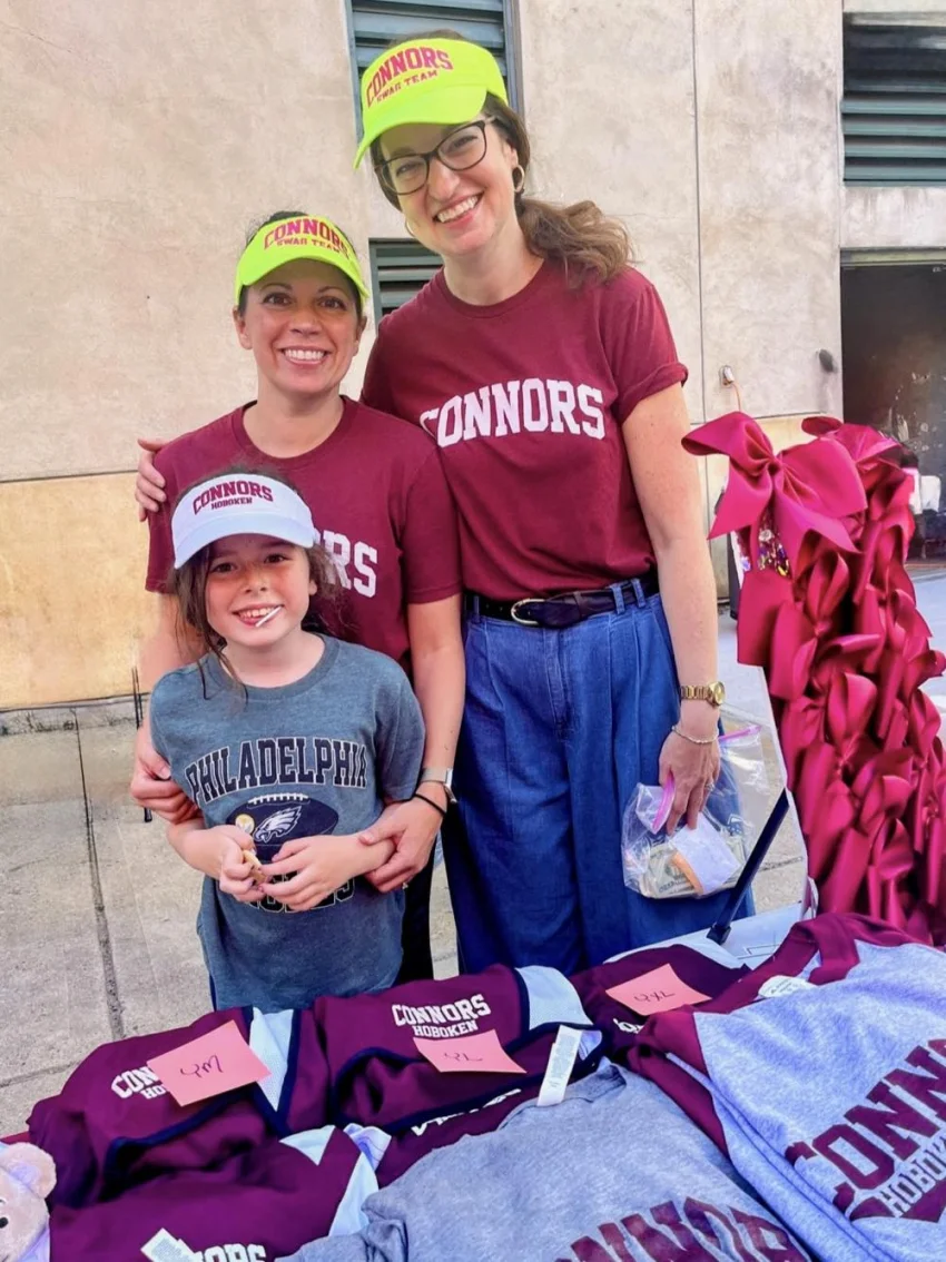 Two Connors Swag Team members in maroon Connors tees and neon Swag Team visors, with a student, behind a table of Connors tees