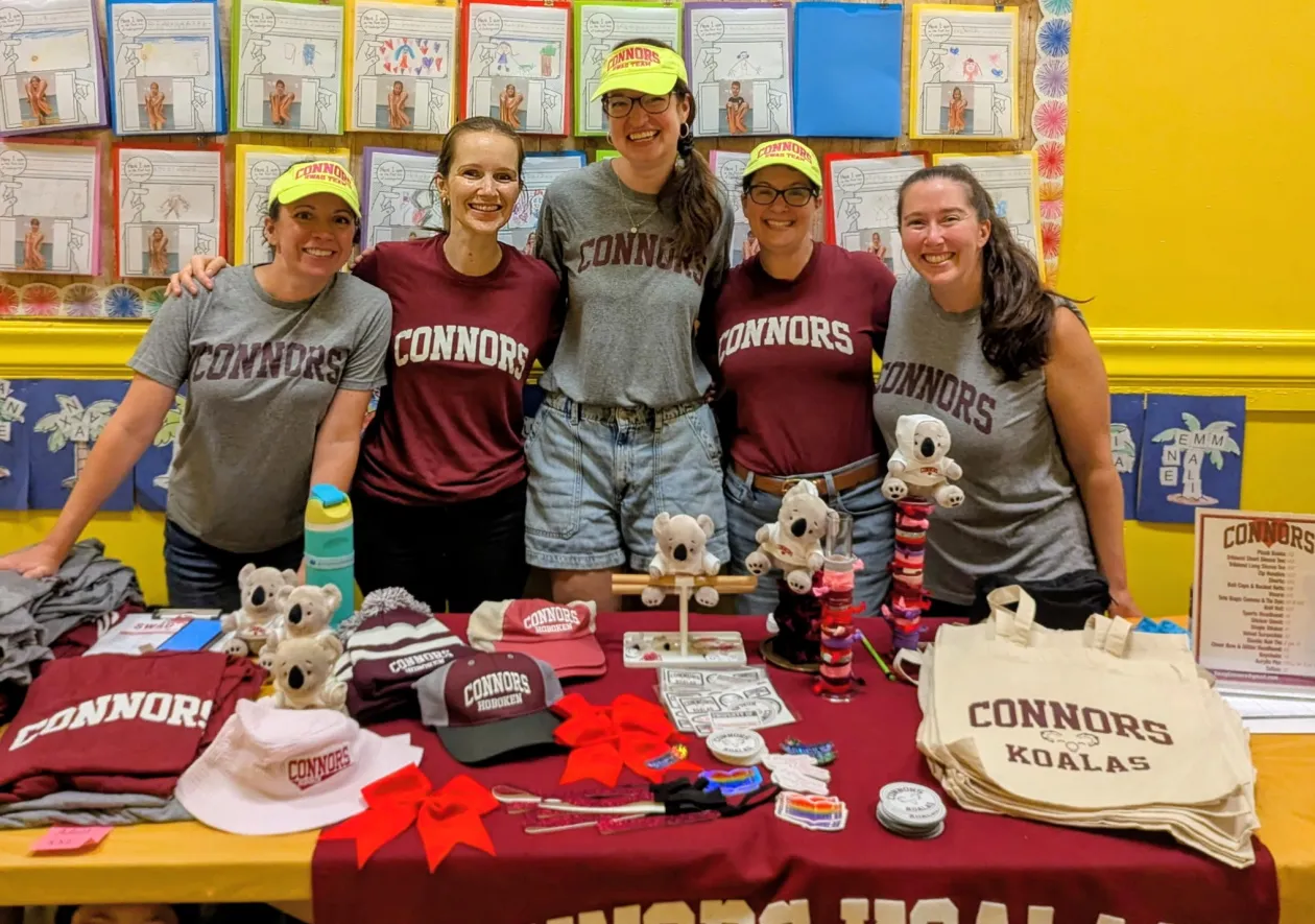 Five Connors Swag Team members in maroon and grey Connors tees behind a swag table of tees, caps, plush Koalas, and tote bags